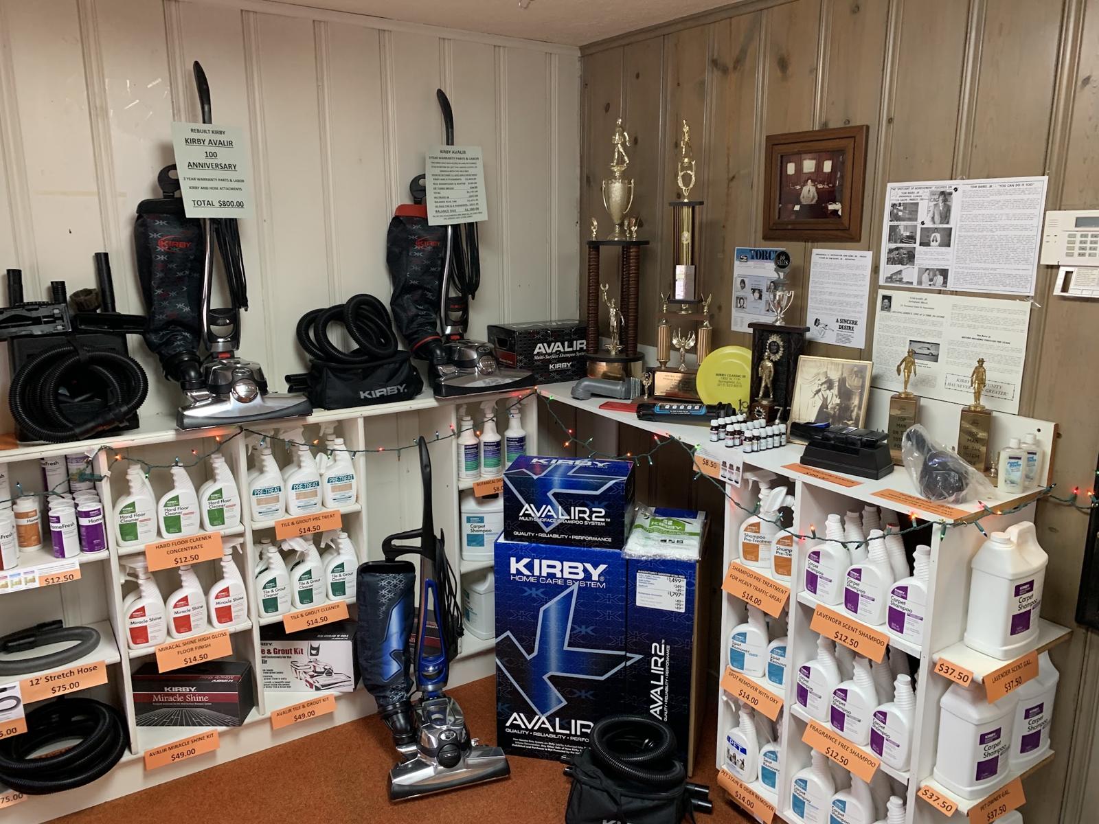 A corner of the showroom with boxed Kirby Avalir 2 systems, hoses, supplies, trophies and framed family history on the wall